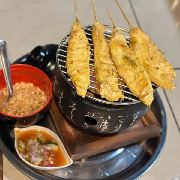 Five Thai chicken satay skewers grilling on a small charcoal grill, with peanut sauce and cucumber relish.