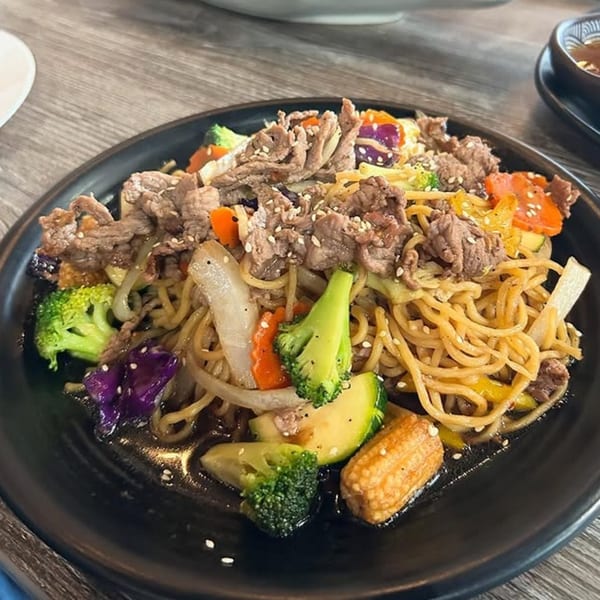 Thai beef stir-fry with noodles, broccoli, carrots, zucchini, and baby corn on a black plate.